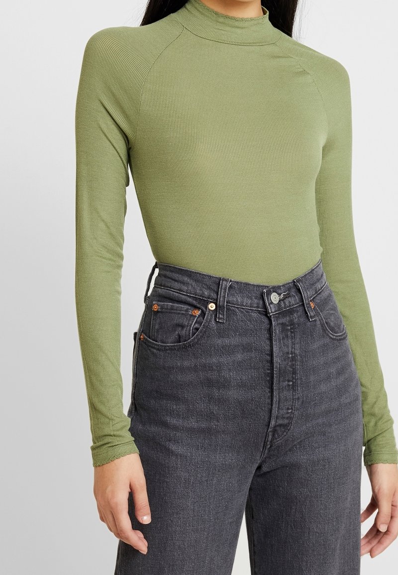 Langärmliges, geripptes Oberteil in Olivgrün mit hohem Kragen, kombiniert mit hochgeschnittenen, dunkelgrauen Jeans mit heller Naht und fünf Taschen.