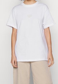 Bílý bavlněný tričko s krátkým rukávem, které na hrudi má malé zlaté logo "JUST DO IT MMXXIII" umístěné na středu. Volný střih.