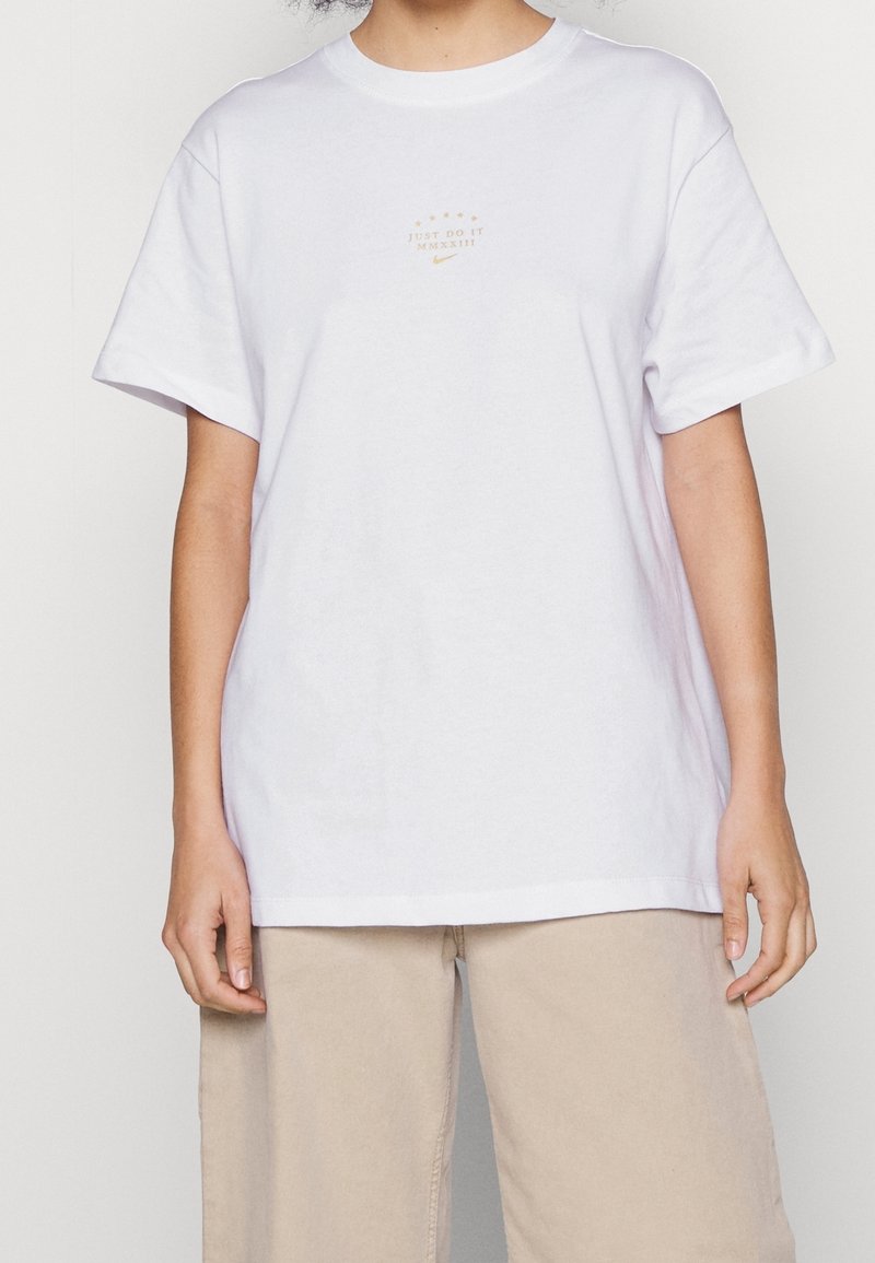 Bílý bavlněný tričko s krátkým rukávem, které na hrudi má malé zlaté logo "JUST DO IT MMXXIII" umístěné na středu. Volný střih.