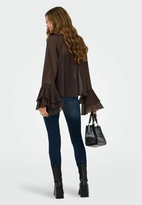 Femme aux cheveux longs et ondulés portant un chemisier marron transparent à manches volantées, un jean skinny foncé, des bottes noires, et tenant un sac à main noir.