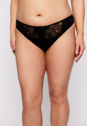 Donna che indossa della biancheria intima nera di pizzo, mostrata dalla parte centrale del torso fino alla parte superiore delle cosce, in piedi contro uno sfondo bianco semplice.