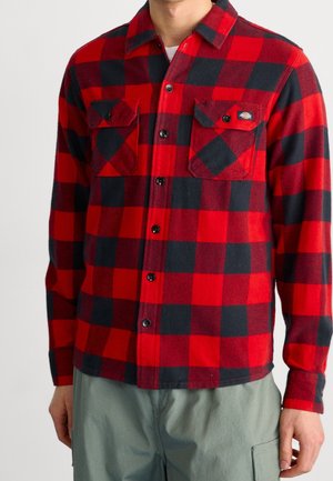 Hombre vistiendo una camisa de franela de cuadros roja y negra con bolsillos en el pecho abotonados y pantalones grises, sobre un fondo liso.