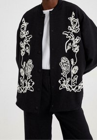 Chaqueta negra con gran bordado floral blanco en la parte delantera, con cuello redondo, cierre de botones y un corte relajado.