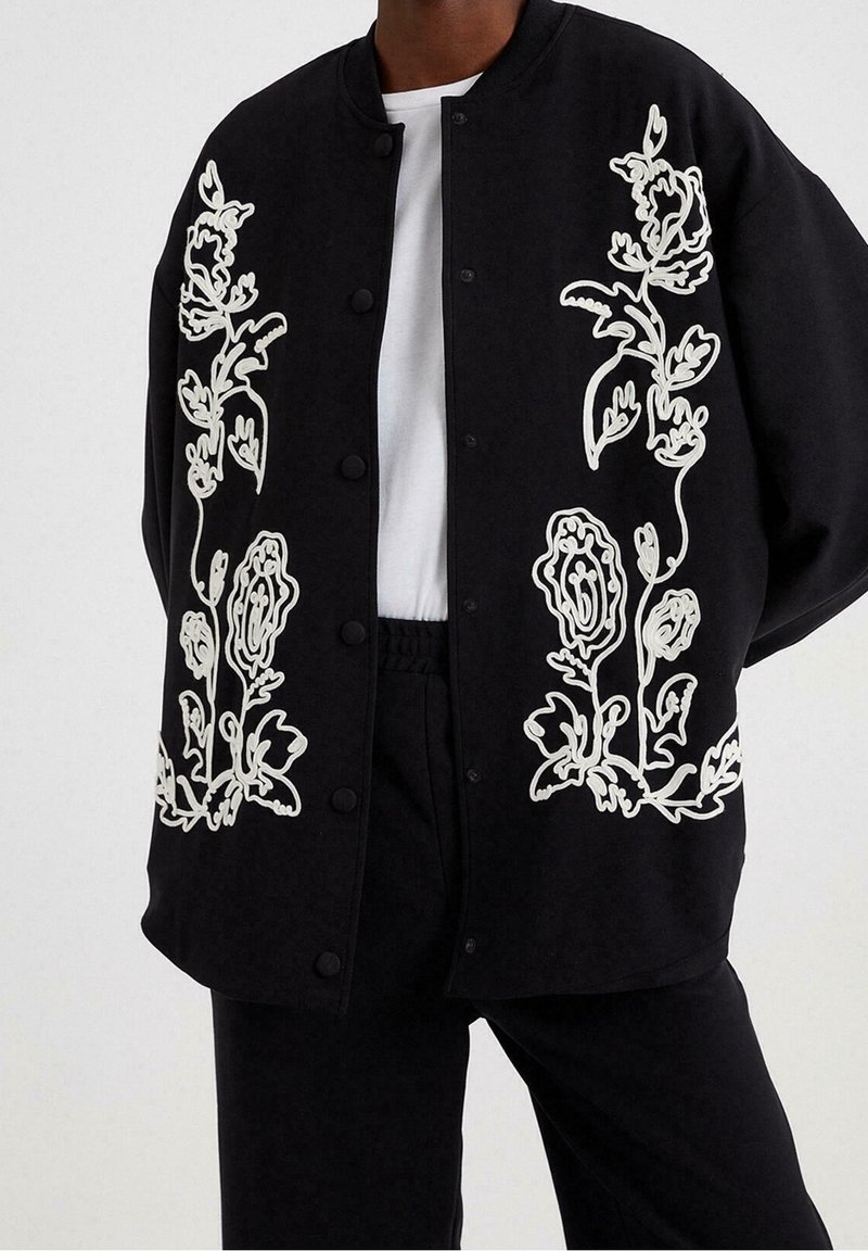 Chaqueta negra con gran bordado floral blanco en la parte delantera, con cuello redondo, cierre de botones y un corte relajado.