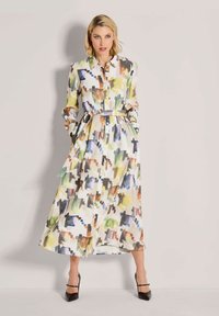 Eine Frau trägt ein langes, mehrfarbiges Shirtkleid mit abstraktem Druck, einem gebundenen Taillengürtel und schwarzen High Heels und steht vor einem schlichten Hintergrund.