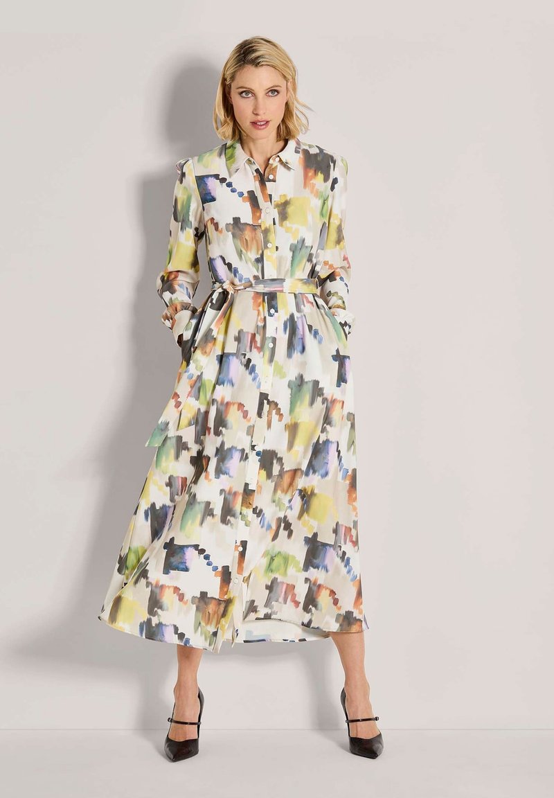 Eine Frau trägt ein langes, mehrfarbiges Shirtkleid mit abstraktem Druck, einem gebundenen Taillengürtel und schwarzen High Heels und steht vor einem schlichten Hintergrund.