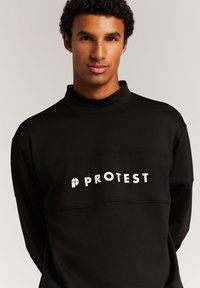 Schwarzer Pullover mit hohem Kragen, der ein weißes "PROTEST"-Logo auf der Vorderseite und kontrastierende Nähte über der Brust aufweist.