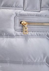 Sekcja jasnoszarej pikowanej kurtki z poziomym przeszyciem. Złoty zamek błyskawiczny z zawieszką w kształcie litery "B". Gładka tekstura.