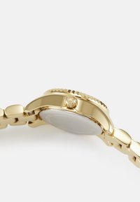 Montre dorée avec un bracelet en acier inoxydable. Boîtier rond, lunette texturée et détail de couronne sur le côté. Surface lisse et finition polie.