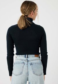 Suéter negro cortado con cuello alto y puños acanalados, combinado con jeans de cintura alta de color azul claro que cuentan con bolsillos traseros y una etiqueta de cuero.