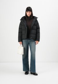 Black puffer jacket with a high collar, paired with a brown turtleneck, flared denim jeans, and a white handbag. Textured black shoes.