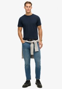 Marineblaues Rundhals-T-Shirt, hellgrauer Pullover um die Taille gebunden, blaue Jeans, schwarzgürtel und schwarze Schuhe in einer lässigen Stehpose.