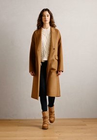Brun ullfrakk med klassisk snitt, over en hvit kabelstrikket genser, kombinert med mørke jeans og brune, pelsforede støvler på et tregulv.