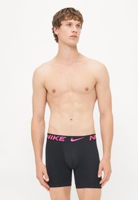 Schwarze Boxershorts mit einem pinken Nike-Logo auf dem Bund. Glattes Material, enganliegendes Design und mittellange Beinlänge bis zur Mitte des Oberschenkels. Keine Muster oder zusätzlichen Akzente.