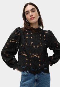 Blusa nera in pizzo con motivo floreale e ritagli. Maniche lunghe a sbuffo e orlo scallop. Abbinata a jeans in denim blu.