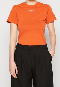 Oranžové tričko s krátkým rukávem a kulatým výstřihem má na hrudi bílý potisk loga "WRSTBHV" a je spojeno s černými kalhotami.
