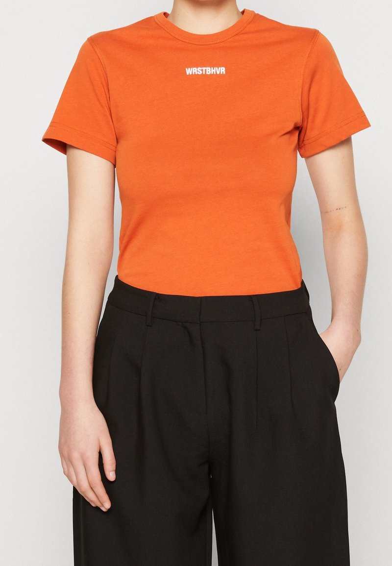 Oranžové tričko s krátkým rukávem a kulatým výstřihem má na hrudi bílý potisk loga "WRSTBHV" a je spojeno s černými kalhotami.