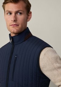 Mörkblå vadderad väst med hög krage och en brun detalj vid axeln. Har en zippered bröstficka och en strukturerad stickad tröja under.