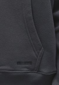 Gros plan d'une poche de sweat-shirt gris foncé avec des coutures visibles et un logo subtil "Hollister" brodé près du bord inférieur.