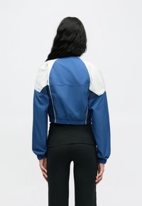 Veste bleue raccourcie avec accents blancs, manches et ourlet élastiques. Texture lisse, design sportif, adaptée pour une tenue décontractée.