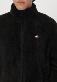 Chaqueta de forro polar negra con cuello alto, cierre de cremallera y un pequeño parche de logo en rojo, blanco y azul en el pecho. Superficie suave y texturizada.