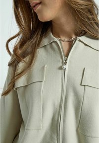 Ljusbeige zip-up jacka med en texturerad finish, som har två bröstfickor, en spetsig krage och en dekorativ dragkedjehållare i pärlor.