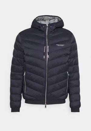 Giacca trapuntata navy con cappuccio, caratterizzata da cuciture a zigzag, dettagli in metallo argentato, tasche con zip e logo sul petto.