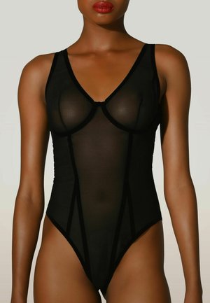 Modelo con un body negro de malla transparente, escote pronunciado en V y copas estructuradas, sobre un fondo liso y claro.