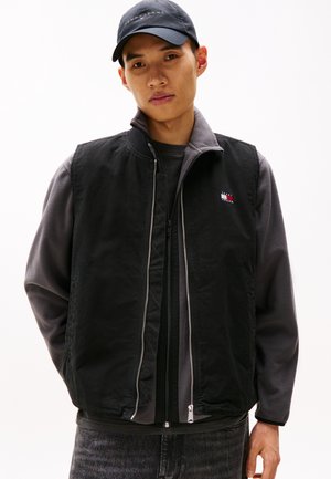 Chaleco sin mangas negro hecho de un material suave, con un forro interior gris, cremallera frontal y un pequeño logo en el pecho.