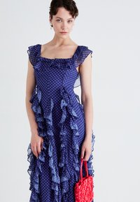 Vestido de lunares en azul marino con acentos de volantes, escote cuadrado y tirantes transparentes; combinado con un bolso rojo texturizado.