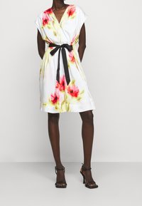 Blommig klänning i vitt tyg med röda och gula blommönster, v-ringning, breda ärmar och ett svart tygbälte i midjan.