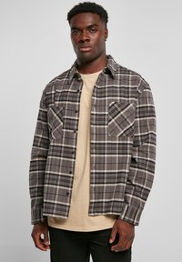 Geruite flanellen overhemd in grijs, zwart en crème met twee borstzakken, knoopsluiting en lange mouwen, gedragen over een beige T-shirt.