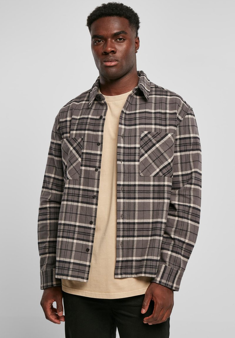 Geruite flanellen overhemd in grijs, zwart en crème met twee borstzakken, knoopsluiting en lange mouwen, gedragen over een beige T-shirt.