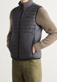 Lätt svart pufferväst med dragkedja fram, horisontell sömnad och sidofickor, lager på en beige tröja.