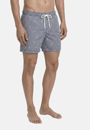 Męski tors i nogi ubrany w granatowo-białe prążkowane kąpielówki z małymi różowymi kotwicami i białym sznurkiem, stojący boso na białym tle.