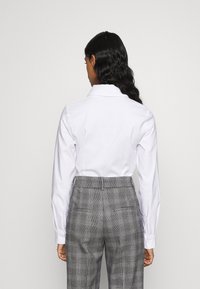 Camicia bianca a maniche lunghe con colletto abbinata a pantaloni grigi a quadri, con vita alta e tasche posteriori, che mostrano una texture liscia.