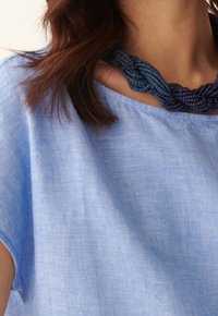 Top de lino azul claro con un escote redondeado, acompañado de un collar de múltiples hebras en tonos azul oscuro y verde azulado, con cuentas texturizadas.