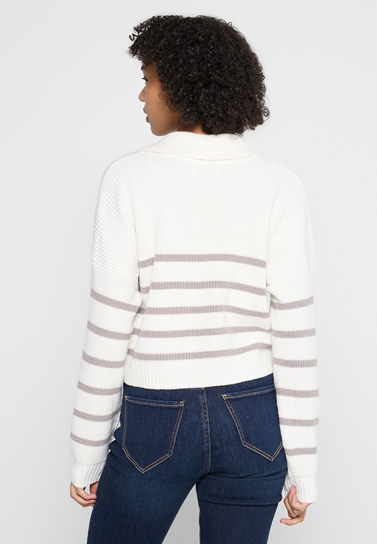 Biały dzianinowy sweter w szare poziome paski, luźny krój i ściągacze na rękawach. W połączeniu z ciemnoniebieskimi jeansami, ukazujący widok z tyłu.