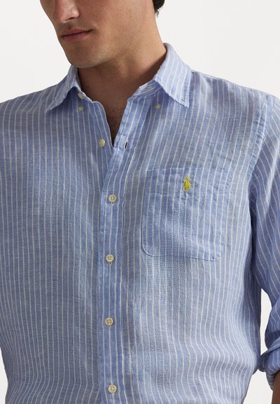 Zila svītraina lina krekls ar krūšu kabatu, pogām lejup, un baltiem pogām. Uz kabatas ir neliels dzeltens emblēma.