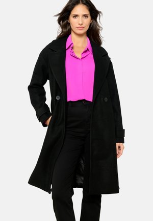 Schwarzer Wollmantel mit breitem Revers und doppelreihigen Knöpfen, über einer leuchtend pinken Bluse getragen, kombiniert mit schwarzen Hosen. Glatte Textur.