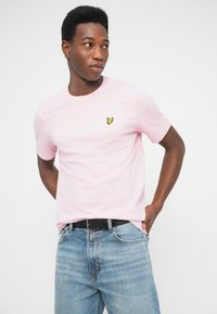 Tricou roz deschis din bumbac, cu guler rotund și mâneci scurte. Are un mic logo galben pe partea stângă a pieptului. Croială casual, fără modele.