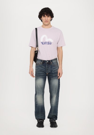 T-shirt violet clair avec un logo graphique bleu, associé à un jean bleu délavé. La personne tient un petit sac noir et porte des chaussures noires.