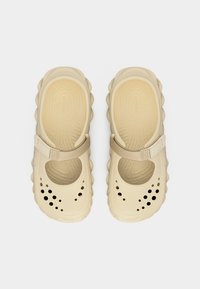 Sabots Crocs beige avec semelle texturée, bride réglable sur le dessus et trous de ventilation circulaires près de la zone des orteils.