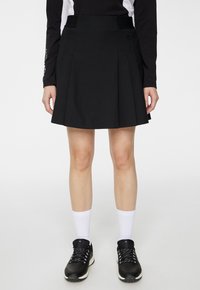 Czarna plisowana spódnica z dopasowaną taliją i subtelnie ukrytymi kieszeniami po bokach, wykonana z lekkiego materiału, wyeksponowana nad białymi sportowymi skarpetkami i czarnymi butami.