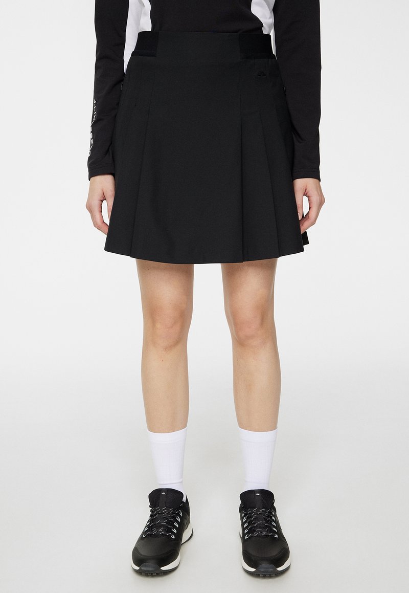 Czarna plisowana spódnica z dopasowaną taliją i subtelnie ukrytymi kieszeniami po bokach, wykonana z lekkiego materiału, wyeksponowana nad białymi sportowymi skarpetkami i czarnymi butami.
