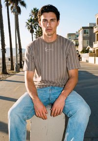 Joven de cabello oscuro, vestido con una camiseta de rayas marrón y jeans azules, sentado sobre un bloque de concreto junto a un paseo marítimo bordeado de palmeras.