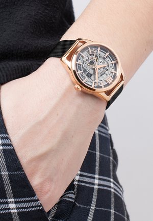 Zapestje z mehansko ročno uro iz rožnatega zlata s črnim mrežastim paščkom, prozornim številčnico, ki razkriva notranje zobnike, in žepom karo hlač.