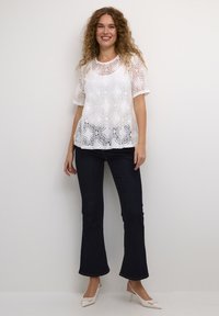 Blusa bianca in pizzo con motivi floreali, maniche corte, abbinata a jeans svasati scuri e tacchi chiari. Tessuto liscio, vestibilità comoda.
