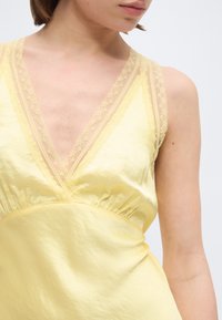 Γυναίκα που φοράει μαν sleeveless κίτρινο σατέν φόρεμα με βαθύ V-λαιμόκοψη διακοσμημένη με δαντέλα, εμφανίζεται από τους ώμους μέχρι τη μέση σε απλό φόντο.