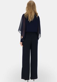 Granatowy kombinezon z zwiewnym, przezroczystym peleryną, szerokimi nogawkami i długim rękawami, z delikatnymi guzikami na mankietach.